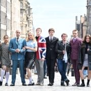 A group of nine young adults stand in a row on a city street, dressed in a mix of retro and modern clothing, channeling Mod Ballet flair, with historic buildings visible in the background.