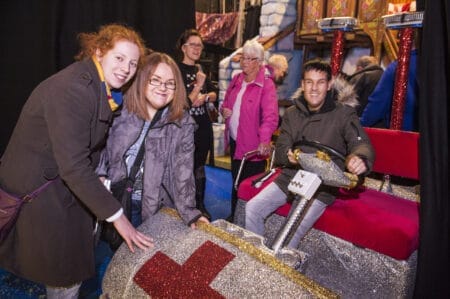 A group of people, some smiling and some standing, interact with a glittery, oversized prop vehicle with a red cross symbol at an indoor event.