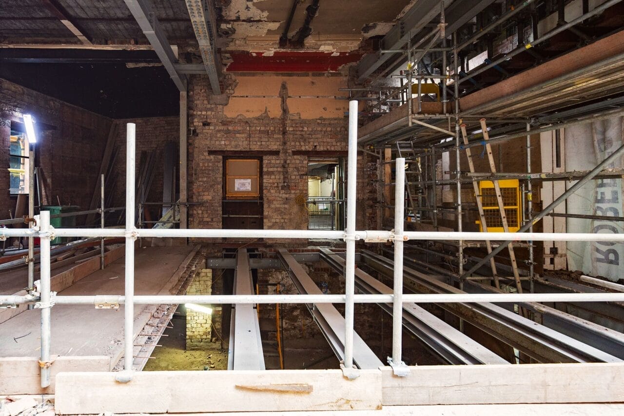 Interior view of the King's Theatre redevelopment, showing exposed brick walls, steel beams, scaffolding, and construction materials. The floor of the cafe area has been dug up and had newly installed steel beams put in.