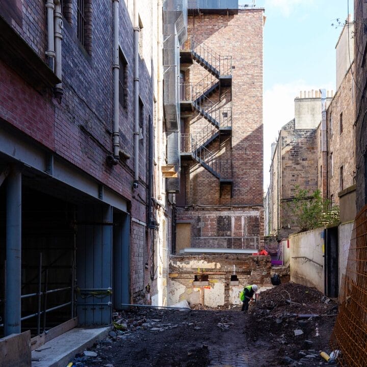 A narrow urban alleyway near King's Theatre under redevelopment, with exposed soil, scaffolding, building materials, and a worker wearing a high-visibility vest making significant strides in construction.