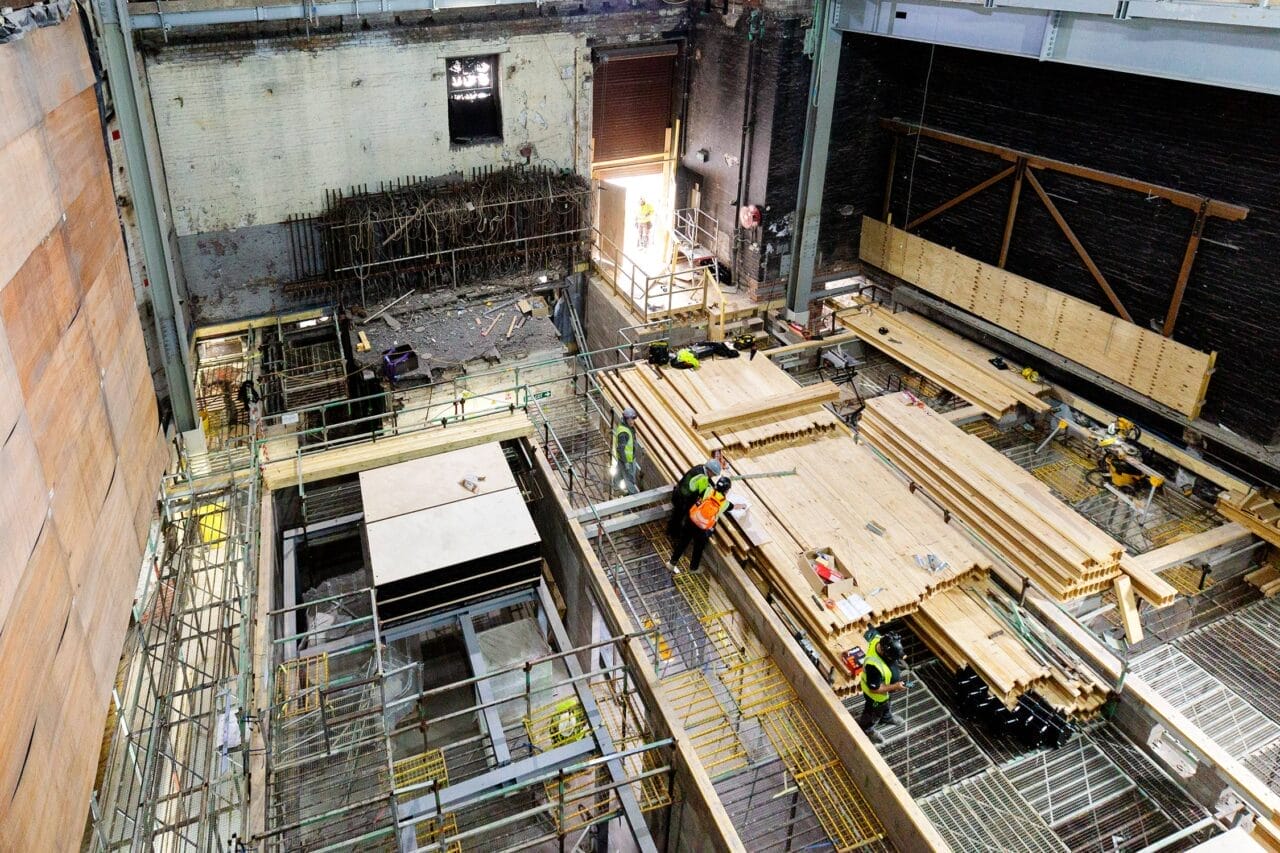 Construction site interior at King's Theatre with workers assembling wooden planks, scaffolding, and building materials scattered. The picture highlights the development of partition walls in the stage basement area.