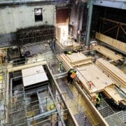 Construction site interior at King's Theatre with workers assembling wooden planks, scaffolding, and building materials scattered. The picture highlights the development of partition walls in the stage basement area.