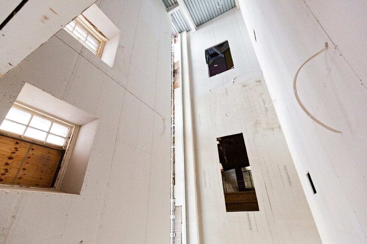 Interior view of a tall, unfinished building shows exposed drywall and hanging electrical wiring with multiple square window openings illuminating the space.