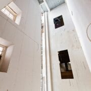 Interior view of a tall, unfinished building shows exposed drywall and hanging electrical wiring with multiple square window openings illuminating the space.