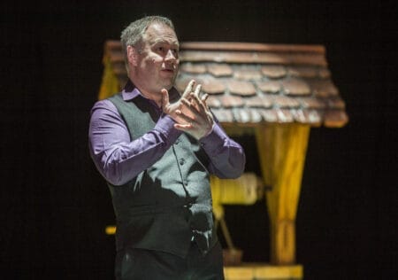 A man in a purple shirt and gray vest gestures with his hands while standing in front of a wooden well on stage.