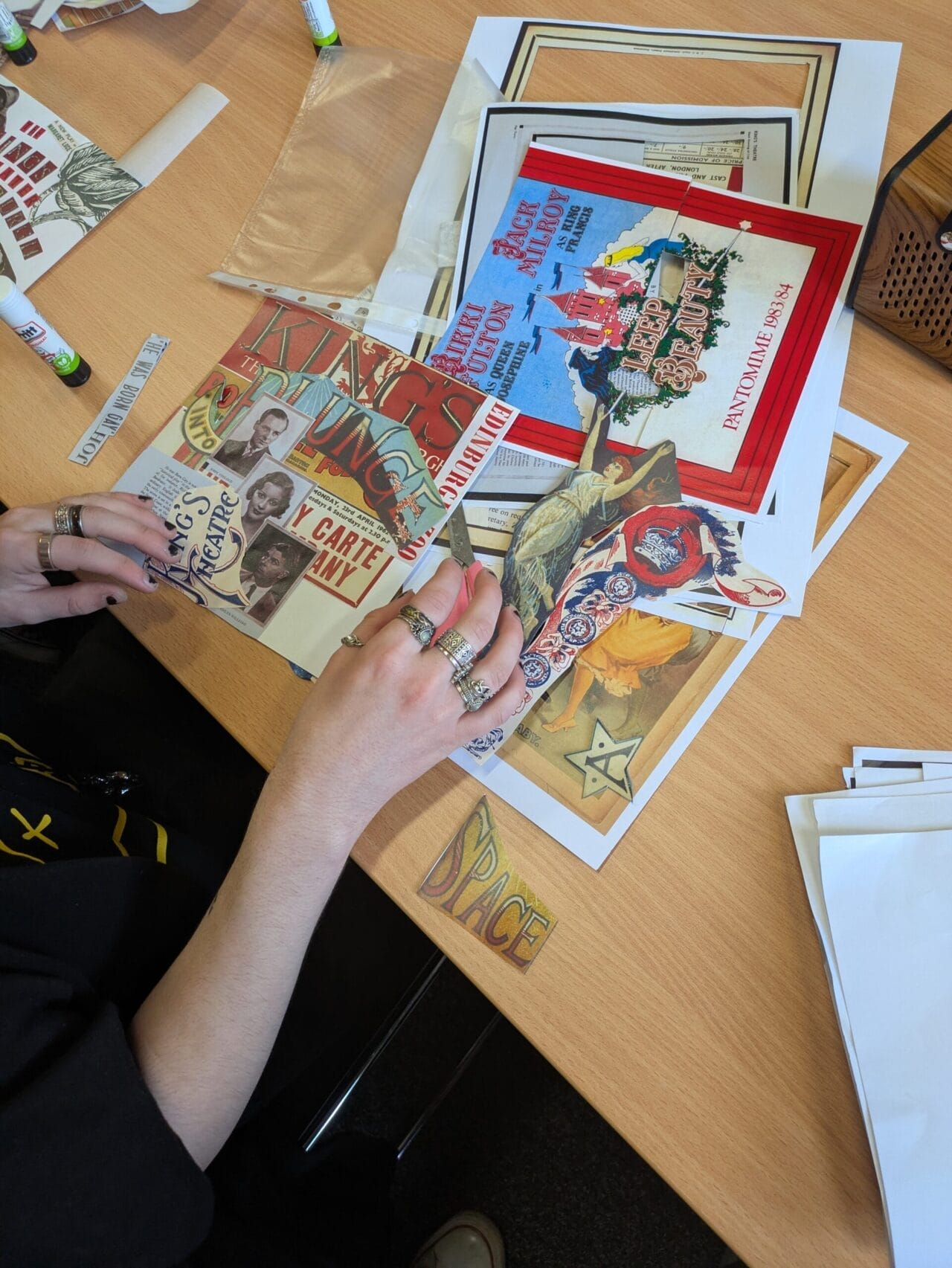 Person arranging vintage-style postcards, photos, and cut-out images on a desk, with glue sticks and other craft supplies, creating a collage inspired by King's Theatre.