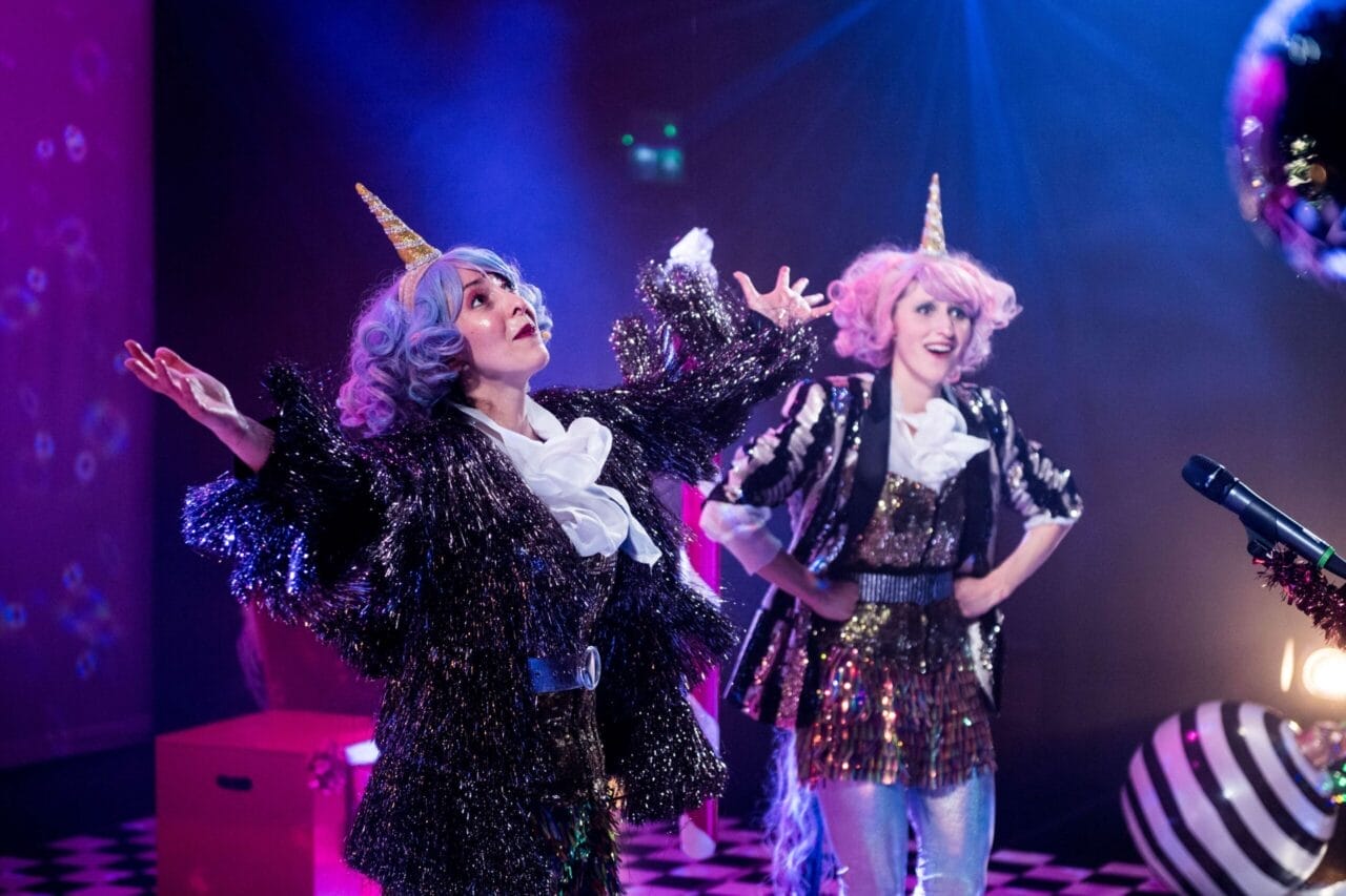 Two performers in colorful wigs, unicorn horn headbands, and sparkly costumes stand on stage under bright lights, as one raises their arms and the other smiles.