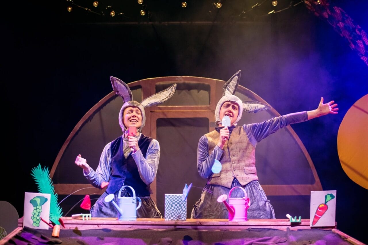 Two people dressed as rabbits with bunny ears perform on stage, holding microphones and surrounded by colorful props.