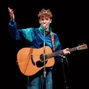 A person wearing a blue jacket and a crown sings while playing an acoustic guitar on stage.