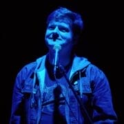 A person stands in front of a microphone, illuminated by blue stage lighting, wearing a denim jacket and looking slightly upward, capturing the anticipation before performing in a new musical theatre work.