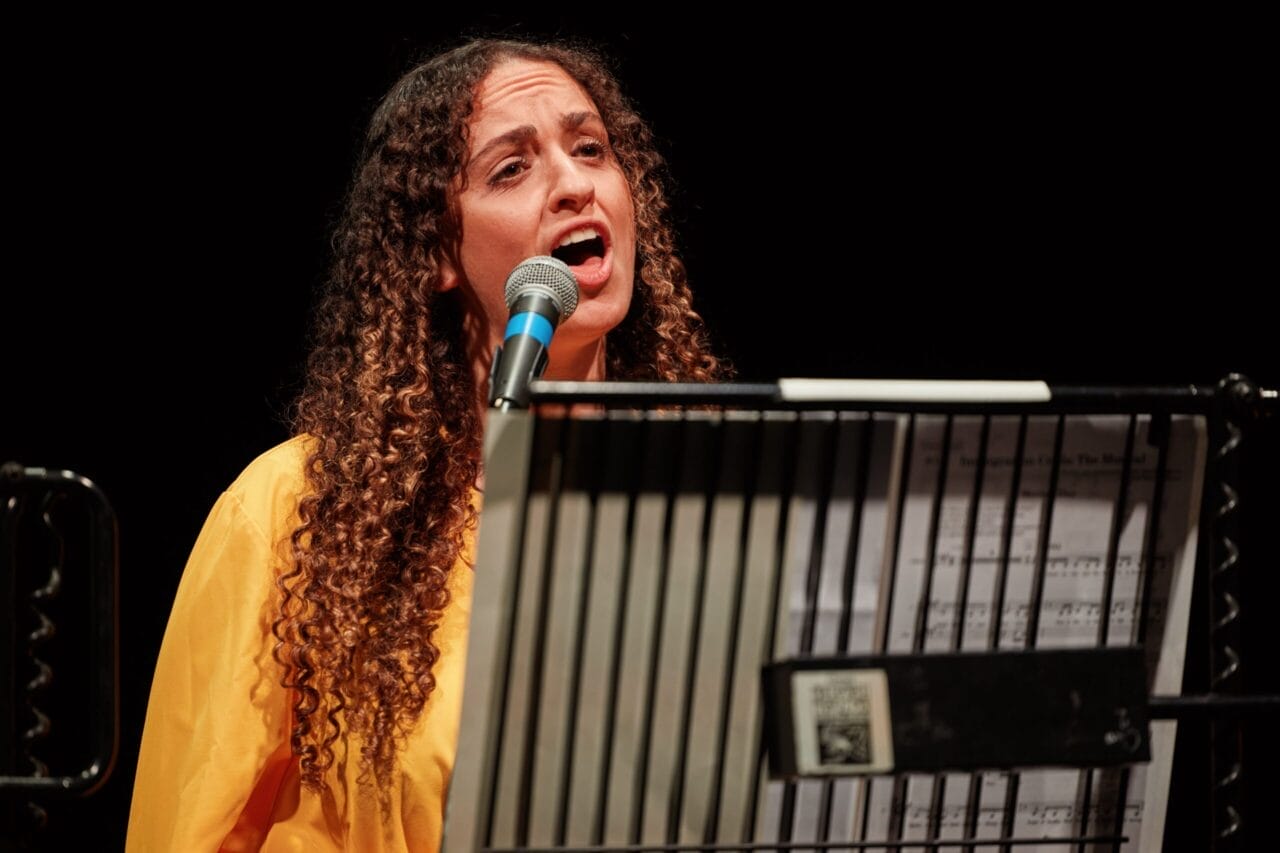 Woman with curly hair in a yellow top sings into a microphone, standing behind a music stand holding sheet music, performing a piece from a new musical.