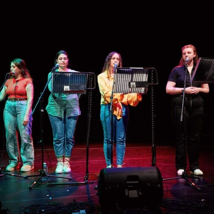 Four women stand on stage in front of music stands and microphones, reading or singing selections from a new musical, illuminated by stage lights against a dark background.
