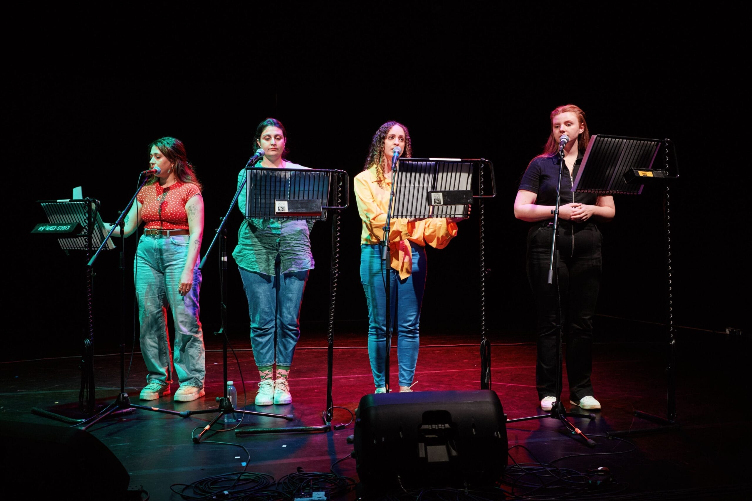 Four women stand on stage in front of music stands and microphones, reading or singing selections from a new musical, illuminated by stage lights against a dark background.
