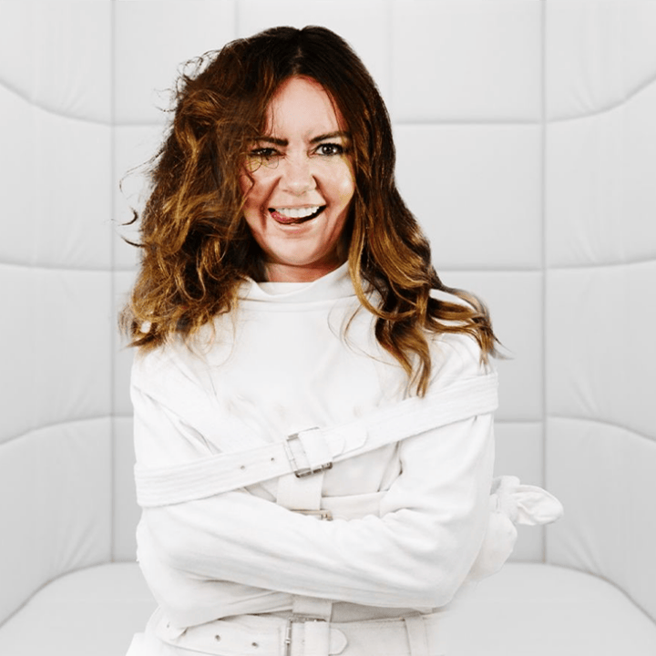 A woman wearing a straitjacket stands in a padded white room, looking directly at the camera with a wild expression, as if awaiting therapy—her intense gaze reminiscent of Serena Terry’s sharp wit.