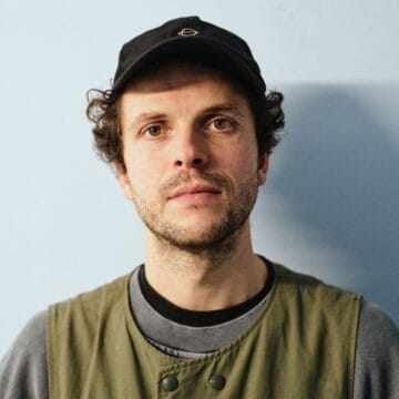 A man with curly hair, wearing a black cap, olive green vest, and gray shirt, stands against a light blue background, looking directly at the camera.