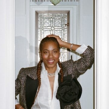 Susan Bender Whitfield stands in a doorway with braided hair, wearing a tweed jacket, white shirt, and jeans. She smiles at the camera, one hand resting on her head.