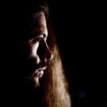 A person with long hair is shown in profile view, partially illuminated against a dark background.