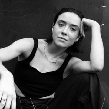 Emma Farnell-Watson, with short dark hair, sits on the ground in a sleeveless black top and dark pants, resting her head on one hand while looking at the camera.