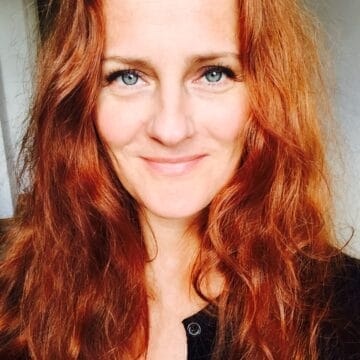 Bregje van Balen, a woman with long, wavy red hair and blue eyes, is smiling at the camera in a close-up portrait. She is wearing a dark top.