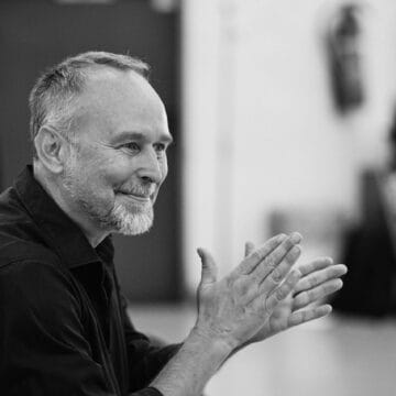 A man with a short beard and mustache smiles and claps his hands while seated indoors. The photo is in black and white.