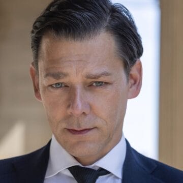 Richard Coyle, dressed in a dark suit and tie, looks directly at the camera with a neutral expression as he stands outdoors near a column.
