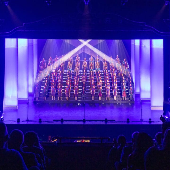A large choir stands on tiered risers, illuminated by stage lights and blue-purple lighting, performing on a theater stage before an audience at the Gang Show Edinburgh.