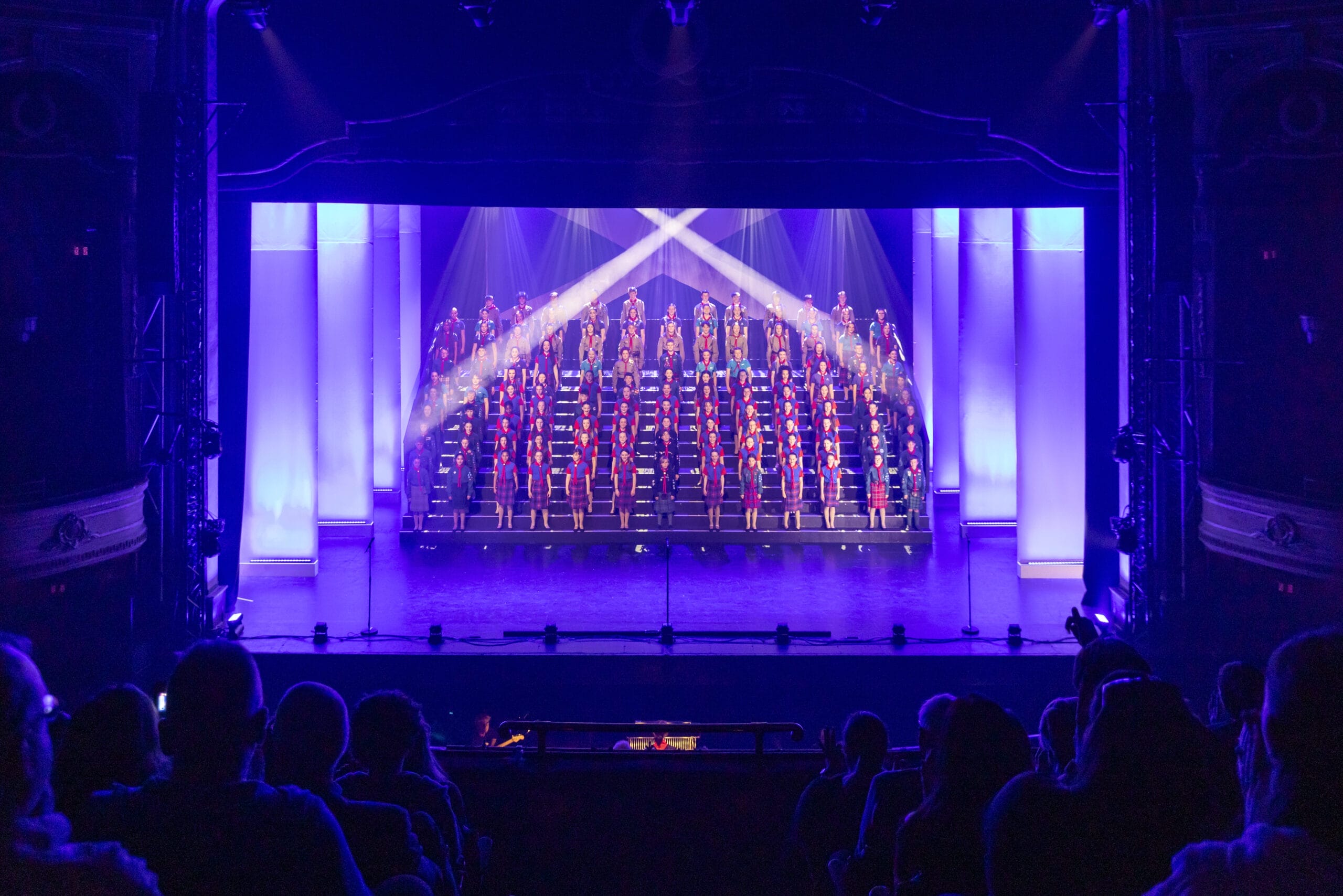 A large choir stands on tiered risers, illuminated by stage lights and blue-purple lighting, performing on a theater stage before an audience at the Gang Show Edinburgh.