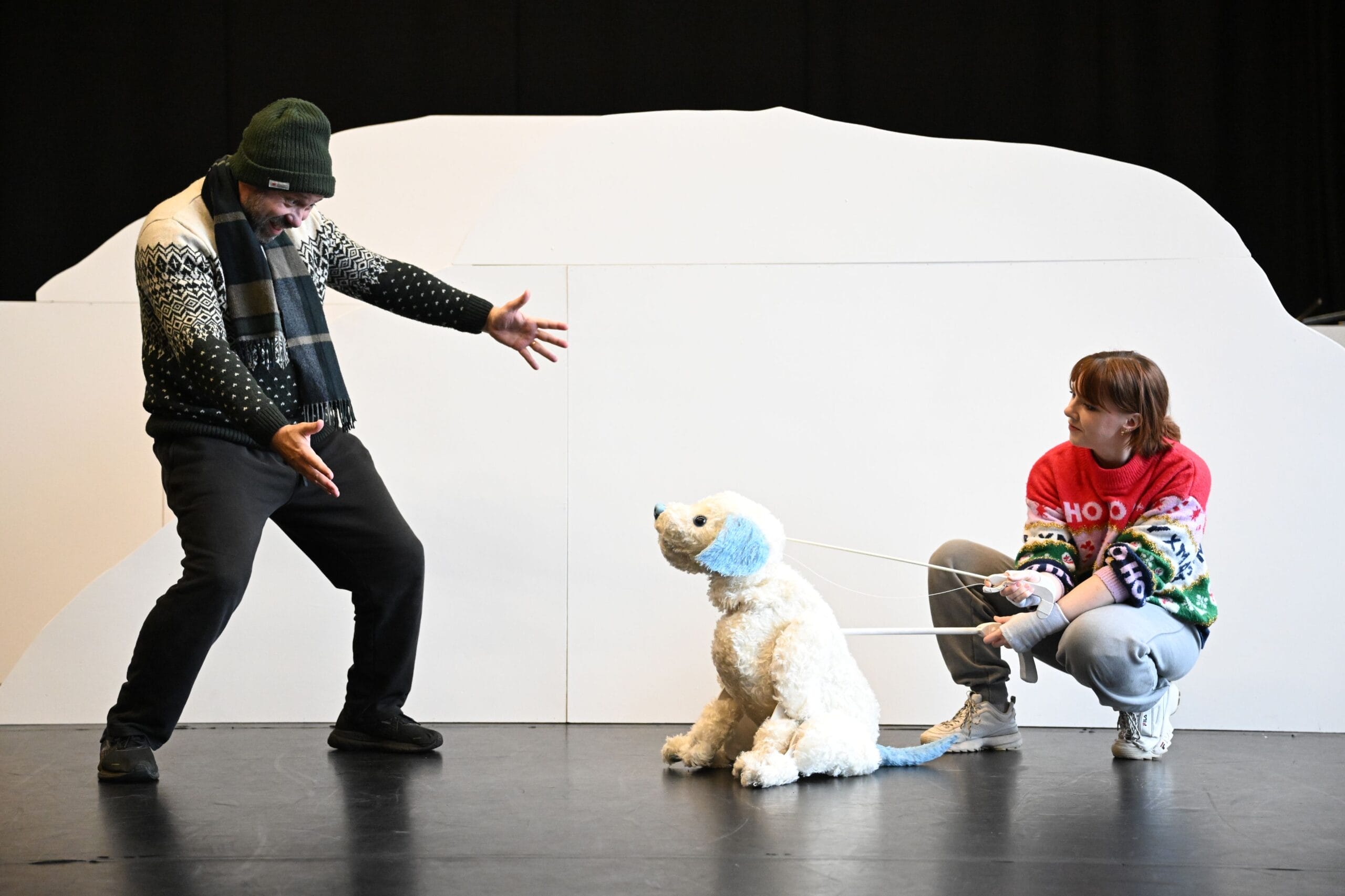 Two people perform on stage with a white polar bear puppet; one person gestures animatedly while the other crouches and holds the puppet’s reins.