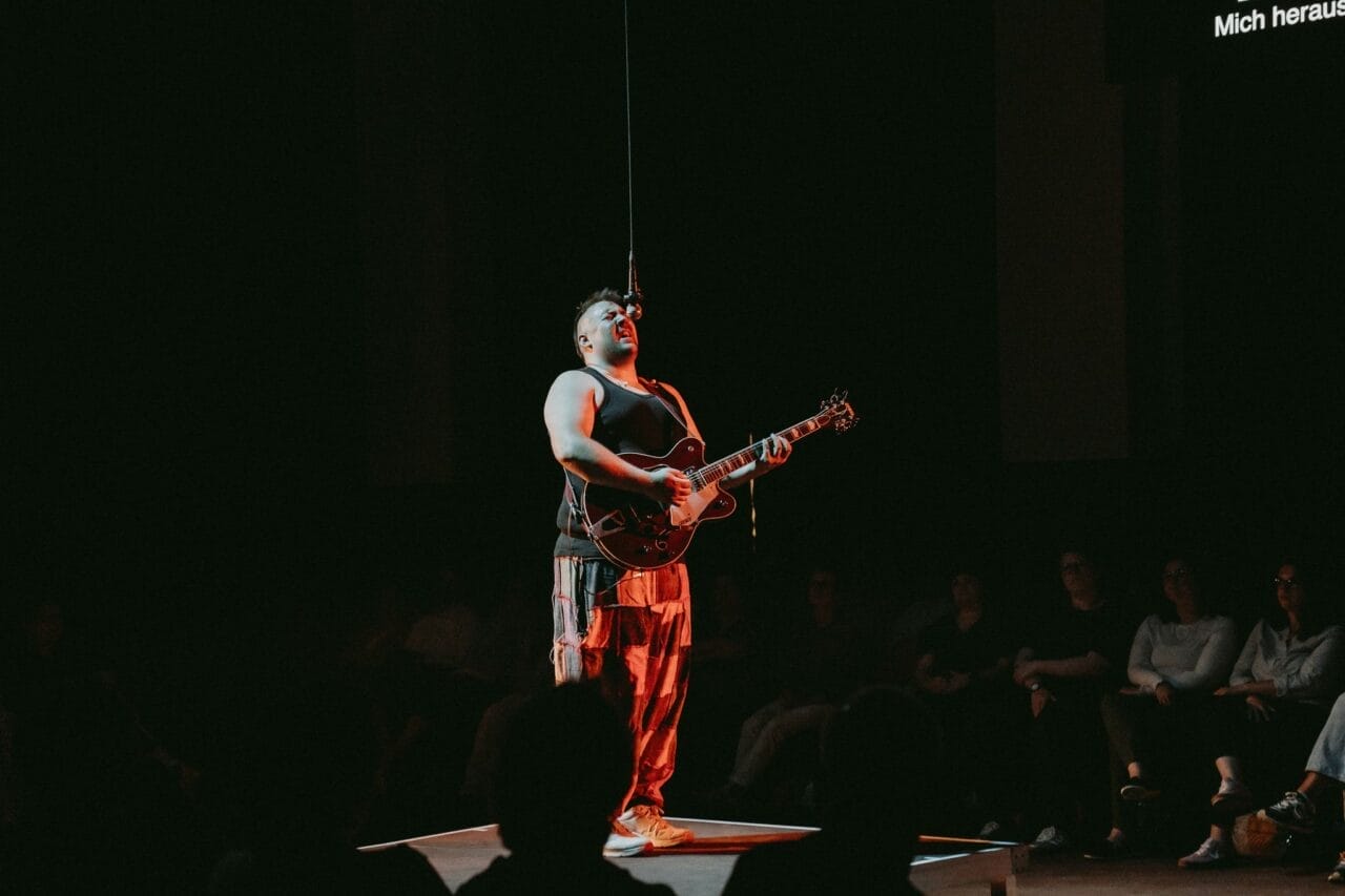 A person stands on a small platform, channeling Half Man Half Bull energy while playing an electric guitar and singing into a microphone, with an audience seated in the background.
