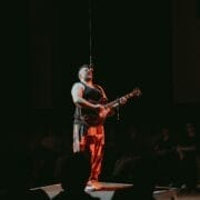 A person stands on a small platform, channeling Half Man Half Bull energy while playing an electric guitar and singing into a microphone, with an audience seated in the background.