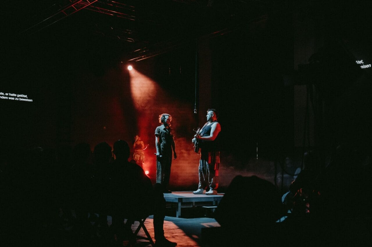Two people stand on a small stage under dramatic lighting, evoking the myth of the Half Man Half Bull; stage lights create a red and dark ambiance as the audience watches from the foreground.