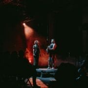 Two people stand on a small stage under dramatic lighting, evoking the myth of the Half Man Half Bull; stage lights create a red and dark ambiance as the audience watches from the foreground.