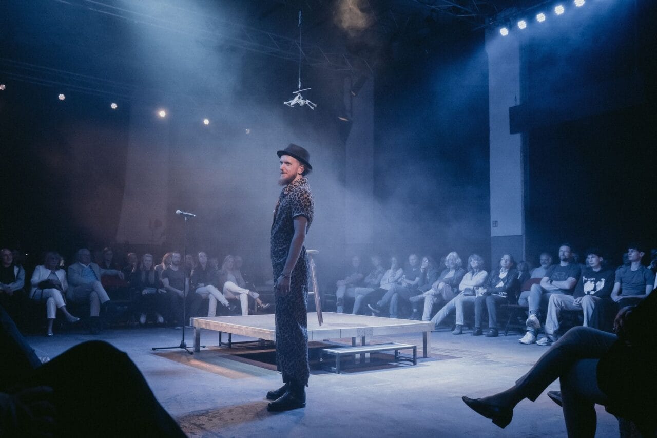 A performer stands on a lit stage in front of an audience seated in a semicircle, with smoke and stage lights creating a dramatic atmosphere, embodying the mythic presence of a Half Man Half Bull.