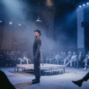 A performer stands on a lit stage in front of an audience seated in a semicircle, with smoke and stage lights creating a dramatic atmosphere, embodying the mythic presence of a Half Man Half Bull.