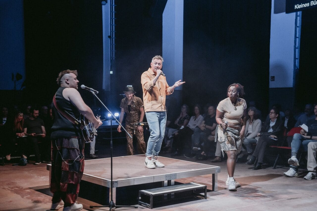 Four performers are on stage in front of an audience; one sings into a microphone while others stand or play guitar under stage lights, evoking the dramatic flair of Greek Mythology and the legendary Minotaur, half man half bull.