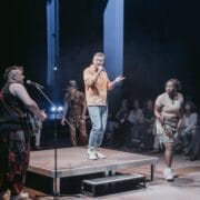 Four performers are on stage in front of an audience; one sings into a microphone while others stand or play guitar under stage lights, evoking the dramatic flair of Greek Mythology and the legendary Minotaur, half man half bull.