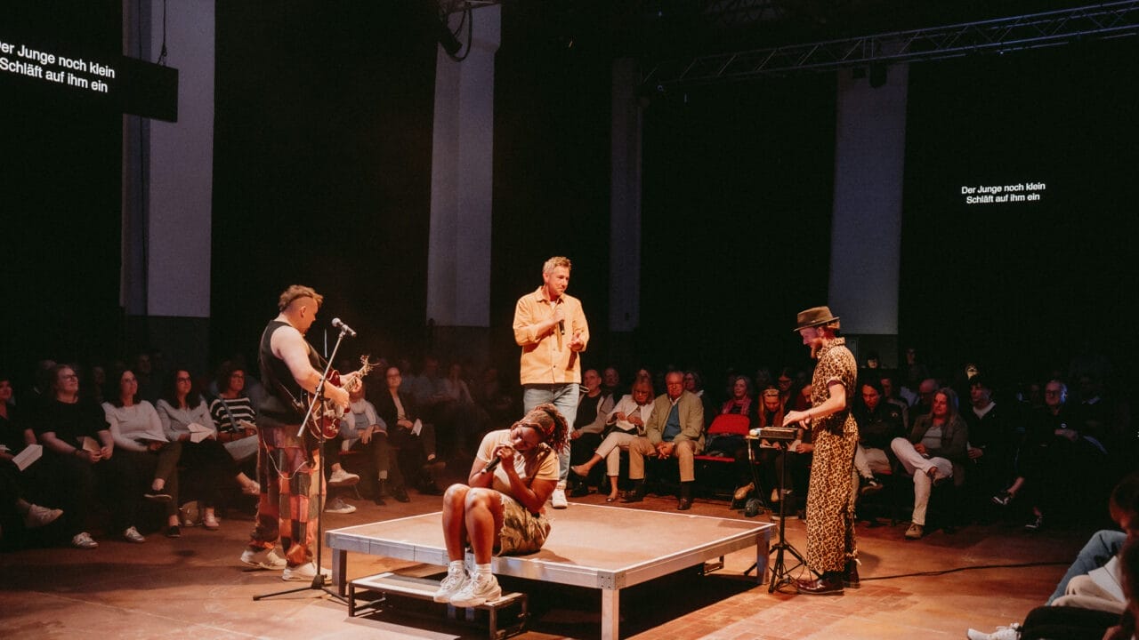 Four performers are on a small stage in front of an audience; two play instruments, one speaks about Half Man Half Bull, and one sits on the platform with their head in their hands.
