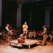Four performers are on a small stage in front of an audience; two play instruments, one speaks about Half Man Half Bull, and one sits on the platform with their head in their hands.