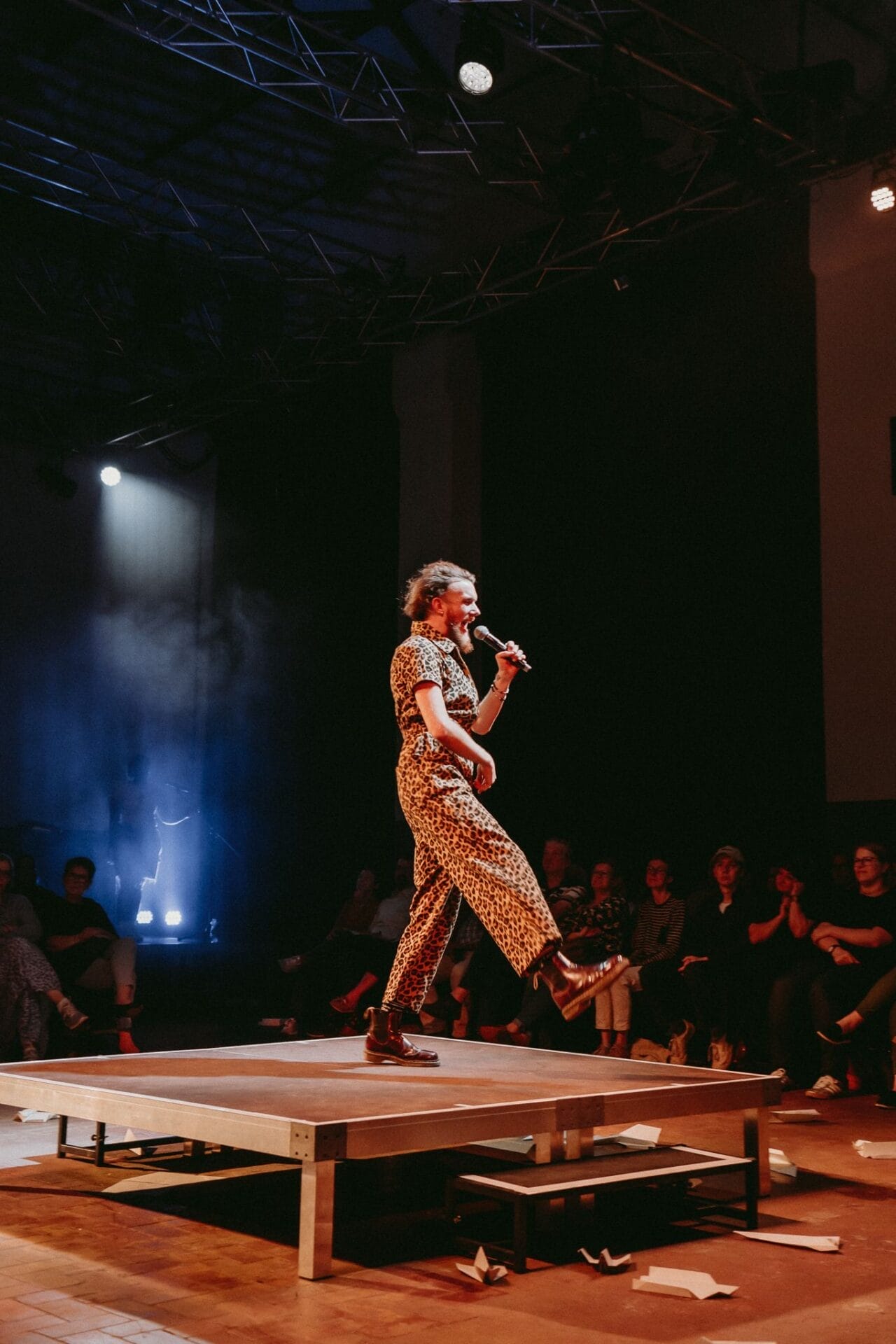 A person in a patterned outfit, channeling a Half Man Half Bull persona, speaks into a microphone while walking on a small platform in front of a seated audience in a dimly lit venue.