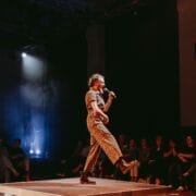 A person in a patterned outfit, channeling a Half Man Half Bull persona, speaks into a microphone while walking on a small platform in front of a seated audience in a dimly lit venue.
