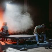 Two people sit back-to-back on a platform under dramatic lighting and smoke, surrounded by paper airplanes, while an audience watches in the background—a scene reminiscent of a modern Half Man Half Bull myth.