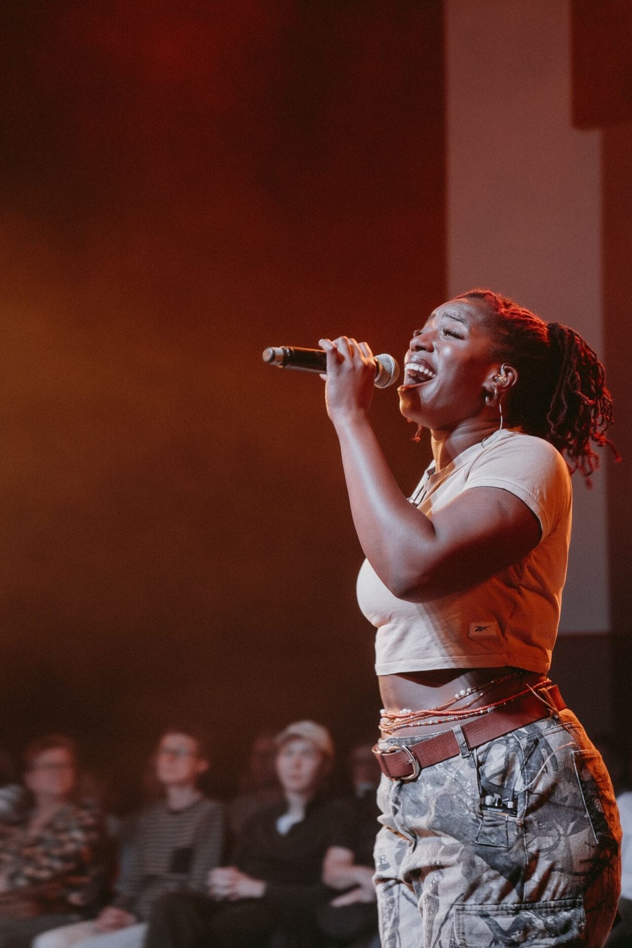 A woman sings passionately into a microphone on stage, channeling Half Man Half Bull energy in her crop top and camouflage pants, with an audience seated in the background.