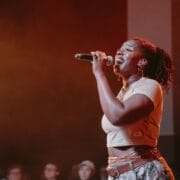 A woman sings passionately into a microphone on stage, channeling Half Man Half Bull energy in her crop top and camouflage pants, with an audience seated in the background.