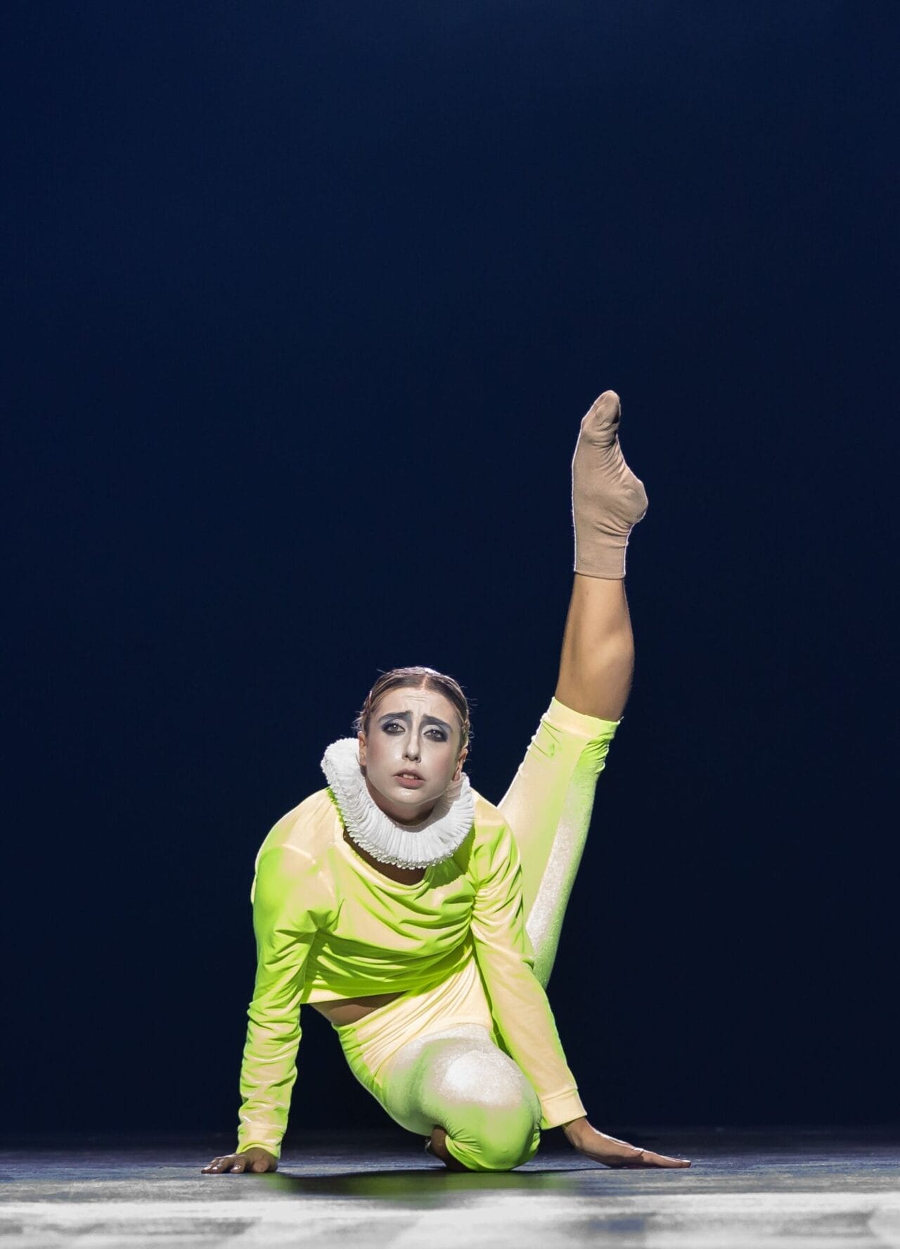 A performer in a neon yellow costume and ruffled collar holds a high leg extension pose on stage under dramatic lighting, evoking the regal flair of Mary Queen of Scots.