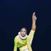 A performer in a neon yellow costume and ruffled collar holds a high leg extension pose on stage under dramatic lighting, evoking the regal flair of Mary Queen of Scots.