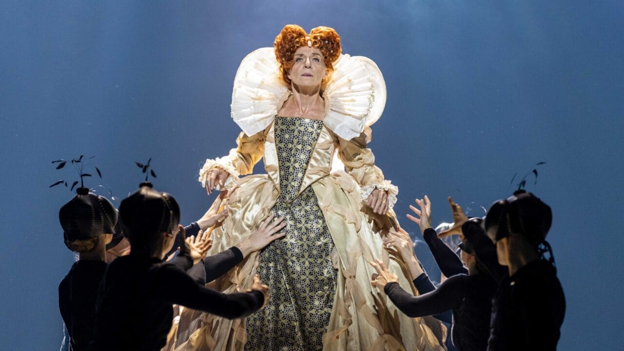 A woman in an elaborate Elizabethan gown stands elevated, evoking Mary, Queen of Scots, while performers in black costumes reach up toward her against a blue-lit background.