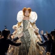 A woman in an elaborate Elizabethan gown stands elevated, evoking Mary, Queen of Scots, while performers in black costumes reach up toward her against a blue-lit background.