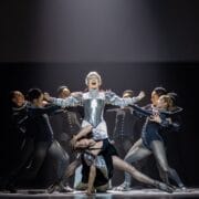 A group of dancers perform on stage, with one central figure in silver armor evoking Mary Stuart, while others pose dynamically around them under dramatic lighting.