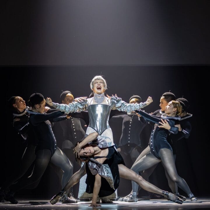 A group of dancers perform on stage, with one central figure in silver armor evoking Mary Stuart, while others pose dynamically around them under dramatic lighting.