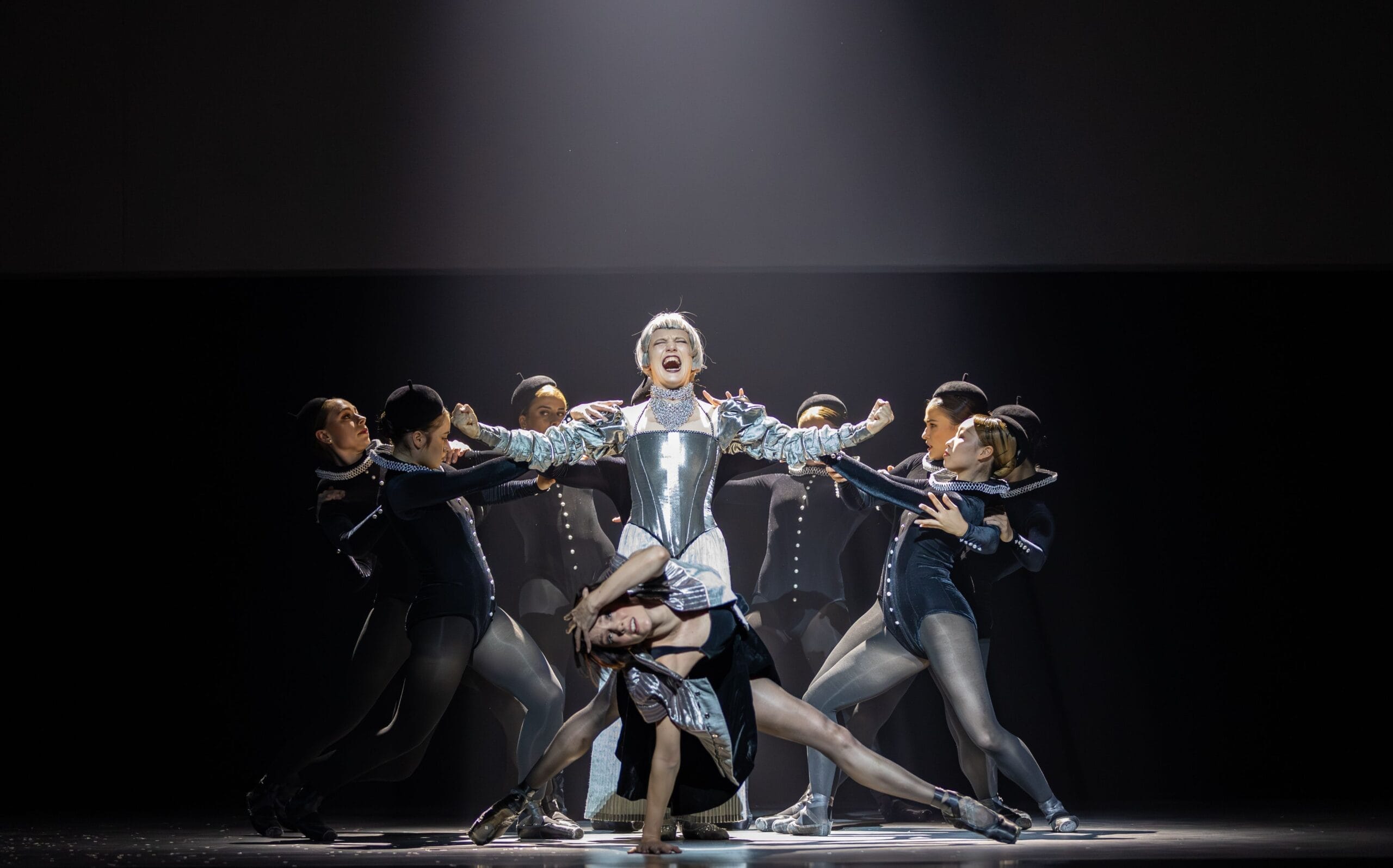 A group of dancers perform on stage, with one central figure in silver armor evoking Mary Stuart, while others pose dynamically around them under dramatic lighting.